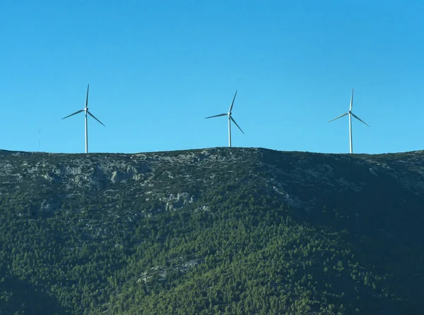 Alternative energy sources concept. Giant white windmills on mountains. Windmills, wind generators on hill, mountain. Landscape with mountains, green forest on sunny day. — Stock Photo, Image