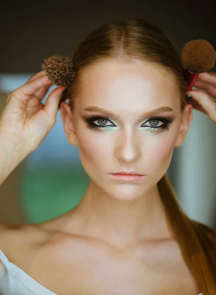 Mujer con cabello rubio y pinceles de maquillaje, belleza. Mujer con maquillaje cara mantenga cepillos en la cabeza. Chica con cara de piel joven, cuidado de la piel, juventud. Modelo de belleza con look glamour, peinado . — Foto de Stock