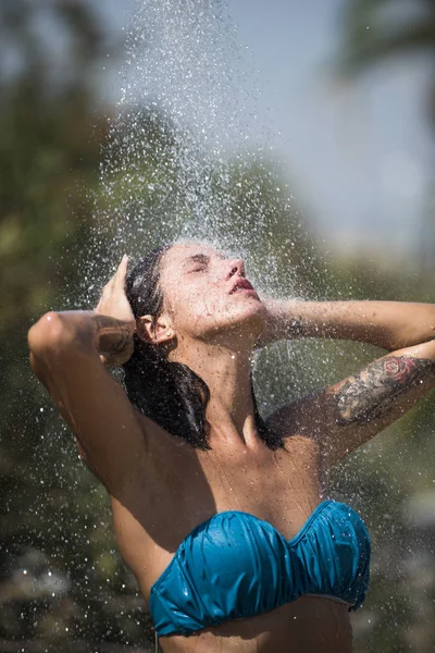 Concepto de calor y refresco. Sexy dama en bikini disfruta de la ducha en el día soleado caliente. Gotas de agua cayendo sobre mujer atractiva. Joven hermosa chica morena tomando ducha en la playa después de nadar . — Foto de Stock