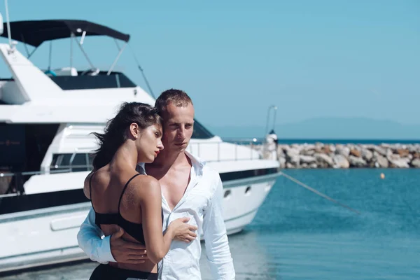 Young couple hugs and relaxing at dock near boat, on sunny summer day. Woman and man in fashionable clothes stands near luxury yacht. Luxury life concept. Couple in love travelling, honeymoon time.