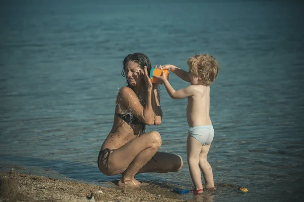 Mom and kid with spend time together at sea beach on sunny day. Cute child boy play with bucket full of water near sea. Family vacation concept. Son pours out water on mother and has fun.