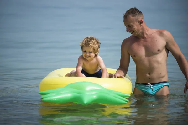 Concepto de paternidad. Padre de pie cerca, empuja el colchón con el hijo puesto. Papá y el niño con caras sonrientes pasan tiempo juntos en el mar. Niño lindo con se sienta en el colchón de aire en forma de piña, en el océano, mar . —  Fotos de Stock