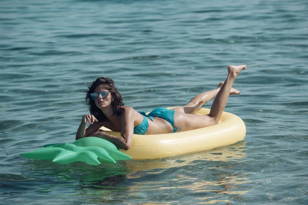 Linda chica nadando en el colchón de aire en forma de piña en el océano, el mar, el uso de bikini elegante y gafas de sol. Concepto de vacaciones y descanso. Mujer tiene descanso, relajarse, tumbarse en el colchón de aire en forma de piña . —  Fotos de Stock