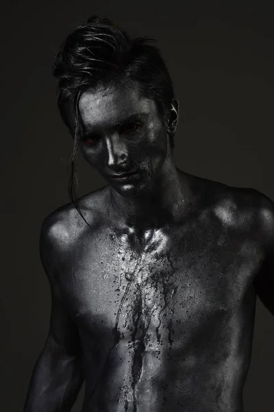 Guy with long hair posing confidently. Athletic figure concept. Macho with streams of sweat or paint on his naked chest. Man with nude torso covered with shimmering silver paint, dark background.