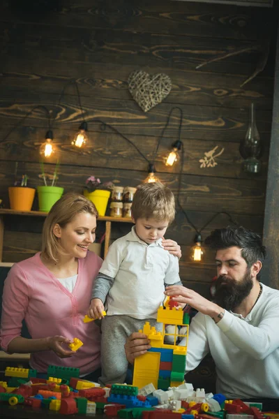 Juego familiar con bloques de plástico juntos. Hermosa mamá sonriente sosteniendo a su lindo hijo por hombro. Rubio niño viendo a su padre construyendo torre de ladrillos de construcción —  Fotos de Stock