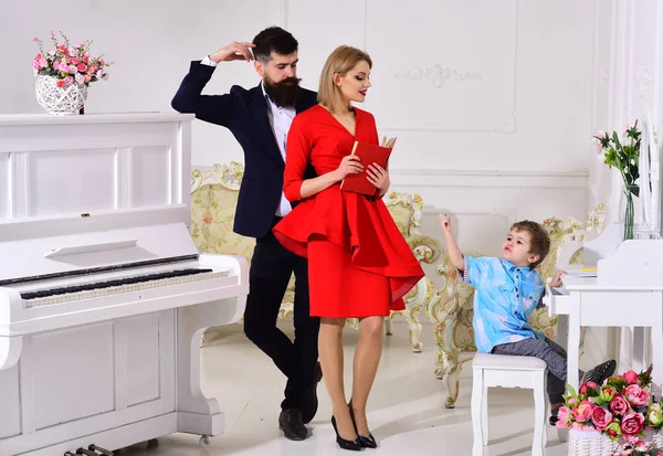 Kid growthing in welfare. Mother and father stands near piano, watching while their son on busy face drawing, luxury interior. Proud parents concept. Parents enjoying parenthood, carefree, happy.