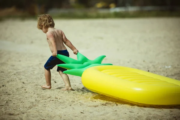 Bambino carino trascinando materasso ad aria in riva al mare. Bambino in vacanza, vacanze estive andando a nuotare sul materasso. Forte ragazzo trascinando materasso a forma di ananas sulla spiaggia di sabbia. Concetto piccolo e forte . — Foto Stock