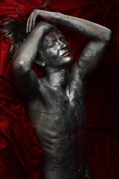 裸の胴体を持つ男は きらめくシルバー塗装 赤い繊維の背景で覆われています スタイリッシュなメイクアップ 不思議な顔の男はシルバー塗装とカラフルな輝くで覆われています ファッションとアートのコンセプト — ストック写真