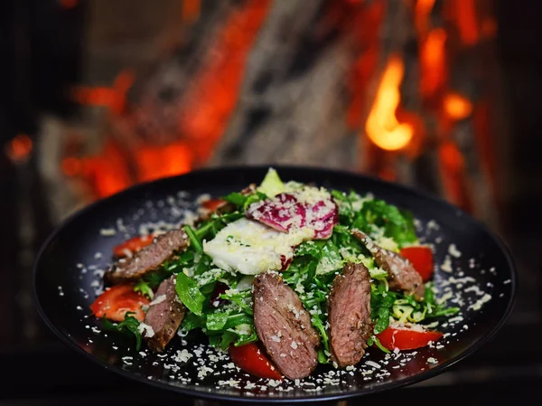 Dish of beef or veal served with lettuce leaves, tomatoes and sprinkled with parmesan cheese, fire on background. Restaurant dish concept. Delicious meat served in restaurant on black dishes.