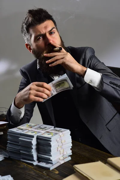 Man sitter vid bord med högar av pengar, räkna vinst. Olagliga kontanter koncept. Mannen i kostym röka cigarr medan räknar pengar. Affärsman på allvarliga ansikte innehar pengar, grå bakgrund. — Stockfoto