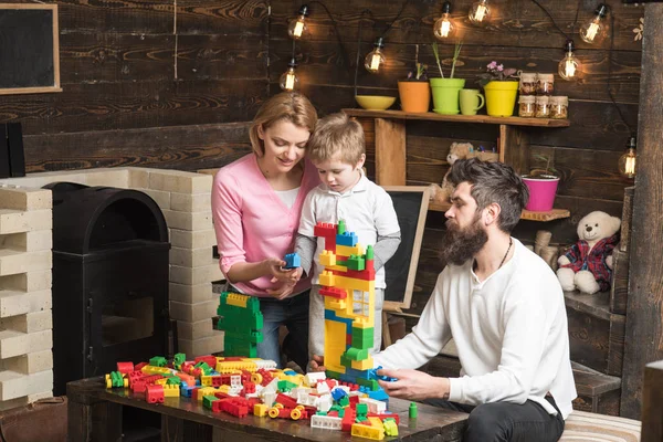 Mamá, papá y niño sentados alrededor de la mesa con ladrillos de construcción de colores. Construcción familiar de una casa de bloques de plástico. Madre e hijo eligen piezas para construir una torre de juguete — Foto de Stock