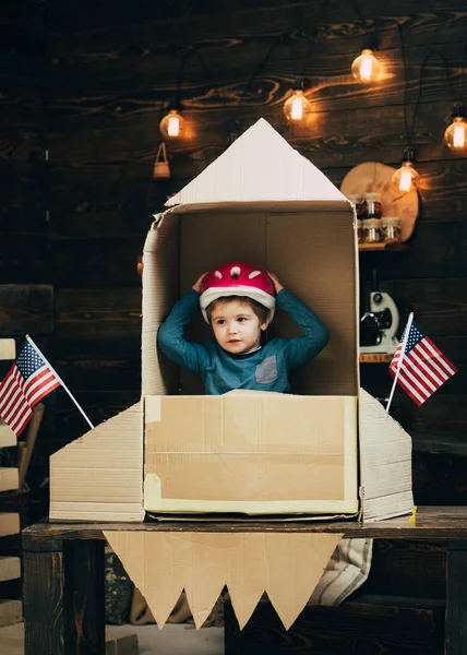 Niño jugar con cohete, cosmonauta sentarse en EE.UU. cohete hecho de caja de cartón. Niño en casco sentarse en cartón cohete hecho a mano con banderas de EE.UU.. Niño jugar astronauta americano. Concepto de cosmonauta . —  Fotos de Stock