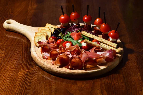 Board with snacks on wooden table. Restaurant dish concept. Cold appetizers with tomatoes, sausage, salami, ham, arugula, olives and bread. Snacks appetizing served on round board. — Stock Photo, Image
