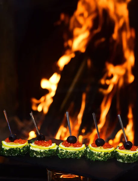 Cold appetizers, canape with red caviar, fire on background. Canape with olives, red caviar, lemon and dill. Restaurant dish concept. Delicious snacks served in restaurant on black dishes. — Stock Photo, Image