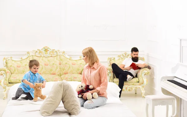 Father not pay attention to kid. Mother play with cute little son, while father busy with book. Indifferent father concept. Mom and son spend time together in bedroom, luxury interior background.