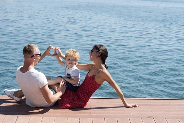 母と父の心や愛を作る彼らの子供に近い手でジェスチャーをします。幸せな家族の時間、一緒に海の背景を過ごします。息子を持つ親は、リアビューで、海辺に座る。家族での休暇の概念. — ストック写真