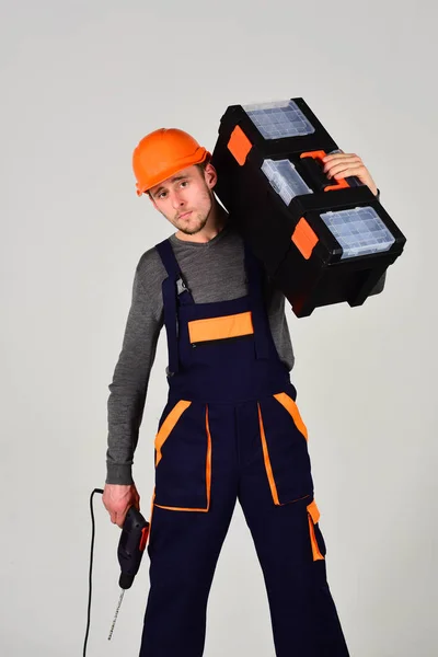 Sterke en aantrekkelijke hersteller, grijze achtergrond, geïsoleerd. Klusjesman concept. Man in overall en helm houdt toolbox op schouder en boor in de hand. Macho op Midden & Zuid Amerika gezicht draagt tools voor reparatie. — Stockfoto