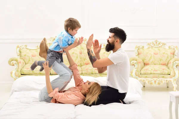 Parents with happy faces pay attention to kid, play, clap hands. Mother and father cuddling with cute son. Young family spend time together on bed, luxury interior background. Happy family concept.