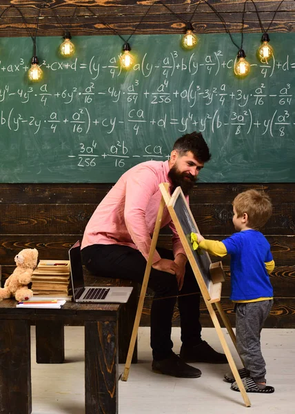 Nachhilfelehrer erklärt dem kleinen Kind Mathe. Junge wischt Kreidetafel mit gelbem Tuch ab. Papa sitzt auf dem Tisch, während er mit dem Sohn spricht — Stockfoto