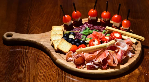 Plateau avec snacks sur table en bois. Snacks appétissants servis sur plateau rond. Concept de plat de restaurant. Entrées froides avec tomates, saucisses, salami, jambon, roquette, olives et pain . — Photo