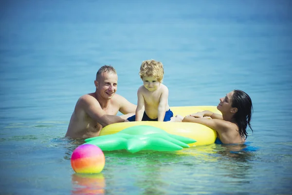 La famille passe du temps ensemble et s'amuse. Mignon enfant garçon assis sur un matelas gonflable en forme d'ananas dans l'océan, la mer, avec les parents. Concept de vacances en famille. Père et mère près du matelas nagent avec leur fils . — Photo