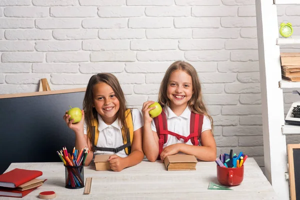 Las niñas comen manzana en la hora del almuerzo. Amistad de las hermanas pequeñas en el aula en el día del conocimiento. Hora de la escuela de chicas. Niños de la escuela feliz en la lección en septiembre 1. Volver a la escuela y a la educación en el hogar — Foto de Stock