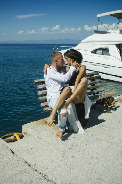 Woman and man in fashionable clothes full of desire near luxury yacht. Couple in love on honeymoon. Honeymoon concept. Young passionate couple sit on bench at dock near boat and kiss, sunny summer day