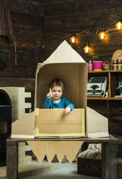 Niño sentado en cartón hecho a mano cohete, apuntando hacia arriba. Chico jugar en casa con cohete, pequeño cosmonauta sentarse en cohete hecho de caja de cartón. Niño jugar cosmonauta, astronauta. Concepto de infancia . —  Fotos de Stock