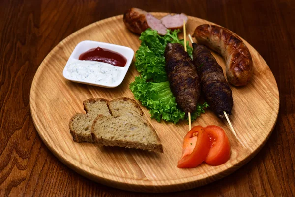 Meat rolls on wooden skewers, shish kebab, grilled sausage served with tomato and sour cream sauce on round wooden board. Restaurant dish concept. Dish appetizing decorated with lettuce and bread.