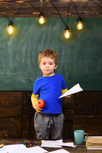 Un petit garçon tenant un avion en papier et une pomme dans ses mains. Écolier en T-shirt bleu debout derrière la table. Concept de jeu éducatif — Photo