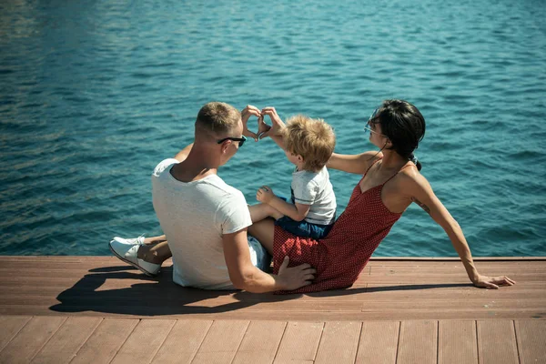 幸福的家庭一起度过时光 海的背景 父母与儿子坐在海滨 后景色 母亲和父亲在他们的孩子附近做心脏或爱手势与手 家庭度假概念 — 图库照片