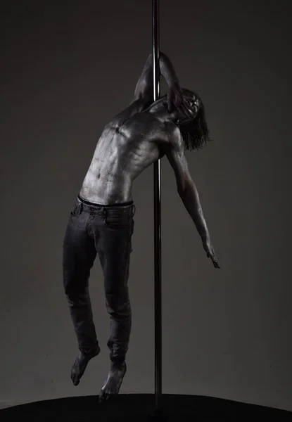 Guy hanging on metallis pole. Performance concept. Man with nude torso covered with shimmering silver paint, dark background. Athlete, sportsman performing pole dancing moves, work out, show trick.