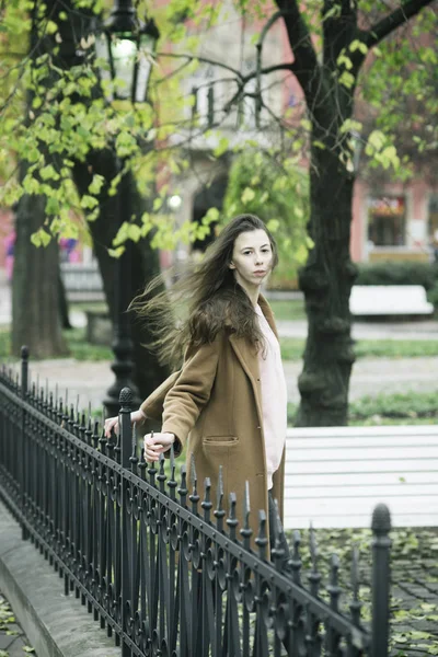 Girl in coat standing outdoors. Woman with calm face waits her boyfriend in park. Windy weather and cozy park. Lady with natural make up and long hair. Beauty, dating, waiting, walk concept.