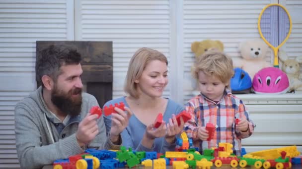 若い家族は 建設プラスチック ブロックでゲームをプレイします 家族ゲームのコンセプト 両親と幸せそうな顔を持つ子供は 赤レンガを保持します 軽い木製の背景にプレイルームで息子と父 — ストック動画