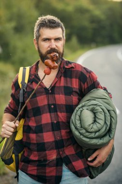 Kasabaya geri yolda otostopçu. Sırt çantası, uyku geri sopa sosis yeme sakallı adam. Ormanda hiking yaz.