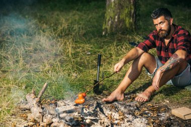 Yalnız gezgin ormanda kamp. Ateşte Sosis kızartma ciddi sakallı adam. Yalınayak hippi orman çim üzerinde oturuyor. Çevre ve doğa kavramı.