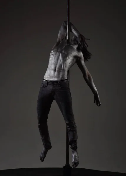 Man with nude torso covered with shimmering silver paint, dark background. Athlete, sportsman performing pole dancing moves, work out, show trick. Guy hanging on metallis pole. Performance concept.