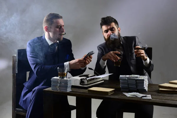 Company engaged in illegal business. Men sitting at table with piles of money and typewriter. Businessmen discussing illegal deal while drinking and smoking, grey background. Illegal business concept.