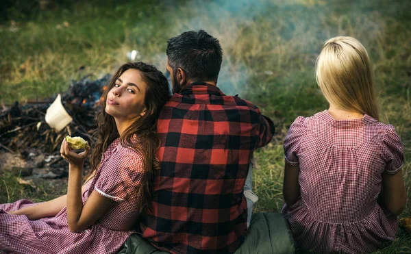 Picnic in the woods. Brunette girl leaning on her boyfriend. Turn back friends sitting on grass next to campfire. Romantic weekend in nature.