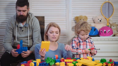 Genç aile inşaat plastik blokları ile oyun oynamak. Aile Oyunları kavramı. Anne ve çocuk mutlu yüzler ile kırmızı tuğla tutun. Baba, anne ve oğlu ışık ahşap arka plan üzerinde oyun odası.