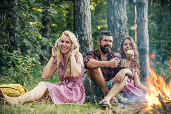 People camping at fire in forest in vintage style. Friends relax at bonfire flame with sparks. Women and bearded man at campfire. Eating food, reading book and entertainment. Summer vacation concept.
