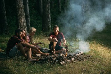 İki çiftler ormanda kamp. Kamp ateşi üzerinde sosis pişirme şık sakallı hippi. Orman sıcak yaz akşamları.