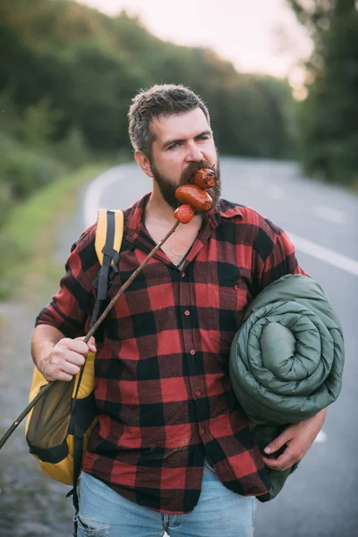 Hitchhiker walking next to highway. Bearded man with rucksack, sleeping back eating sausages from stick. Summer hiking in forest.