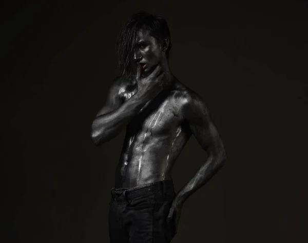 Guy posing with confident sexy expression, holds finger on lips. Macho with streams of sweat on naked chest. Desire concept. Man with nude torso covered with shimmering silver paint, dark background.