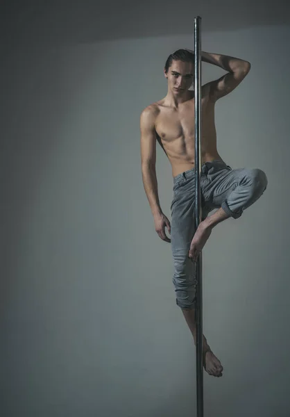 Sport and art concept. Attractive, sexy man with long hair, nude torso, grey background. Guy makes figure on pole, shows trick. Athlete, sportsman performing pole dancing moves, work out, show trick.