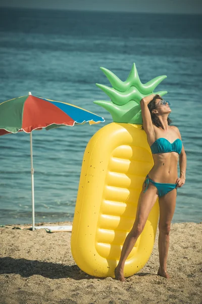 Summer vacation concept. Lady with air mattress pineapple shaped stand at beach, wearing stylish bikini and sunglasses. Woman holds air mattress pineapple shaped with sea or ocean on background.