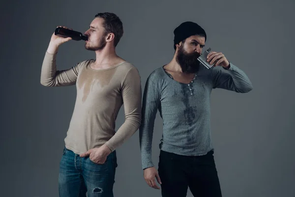 Company of dirty sweaty friends spend leisure with drinks. Alcohol, addiction, leisure. Guys hold bottle and flask with alcohol, drinking. Men on drunk faces, grey background. Alcohol addict concept.