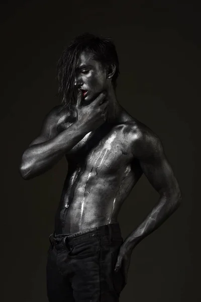 Guy posing with confident sexy expression, holds finger on lips. Macho with streams of sweat on naked chest. Man with nude torso covered with shimmering silver paint, dark background. Desire concept.