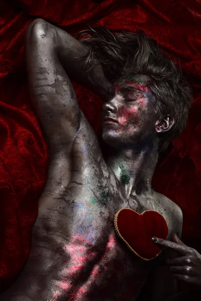 Guy on sad face with make up, covered with shimmering silver paint and colorful glitters. Depression and brake up concept. Man with nude torso holds red plush soft heart toy on chest, dark background.