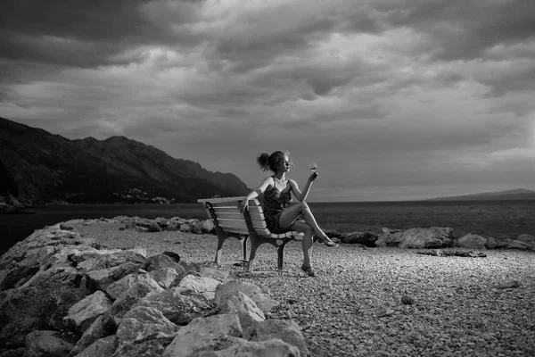 Femme sexy ou fille avec verre de vin se trouve sur le banc en plein air sur un ciel sombre crépusculaire avec des nuages sur la plage avec de l'eau de mer ou de l'eau de mer sur fond naturel du soir — Photo